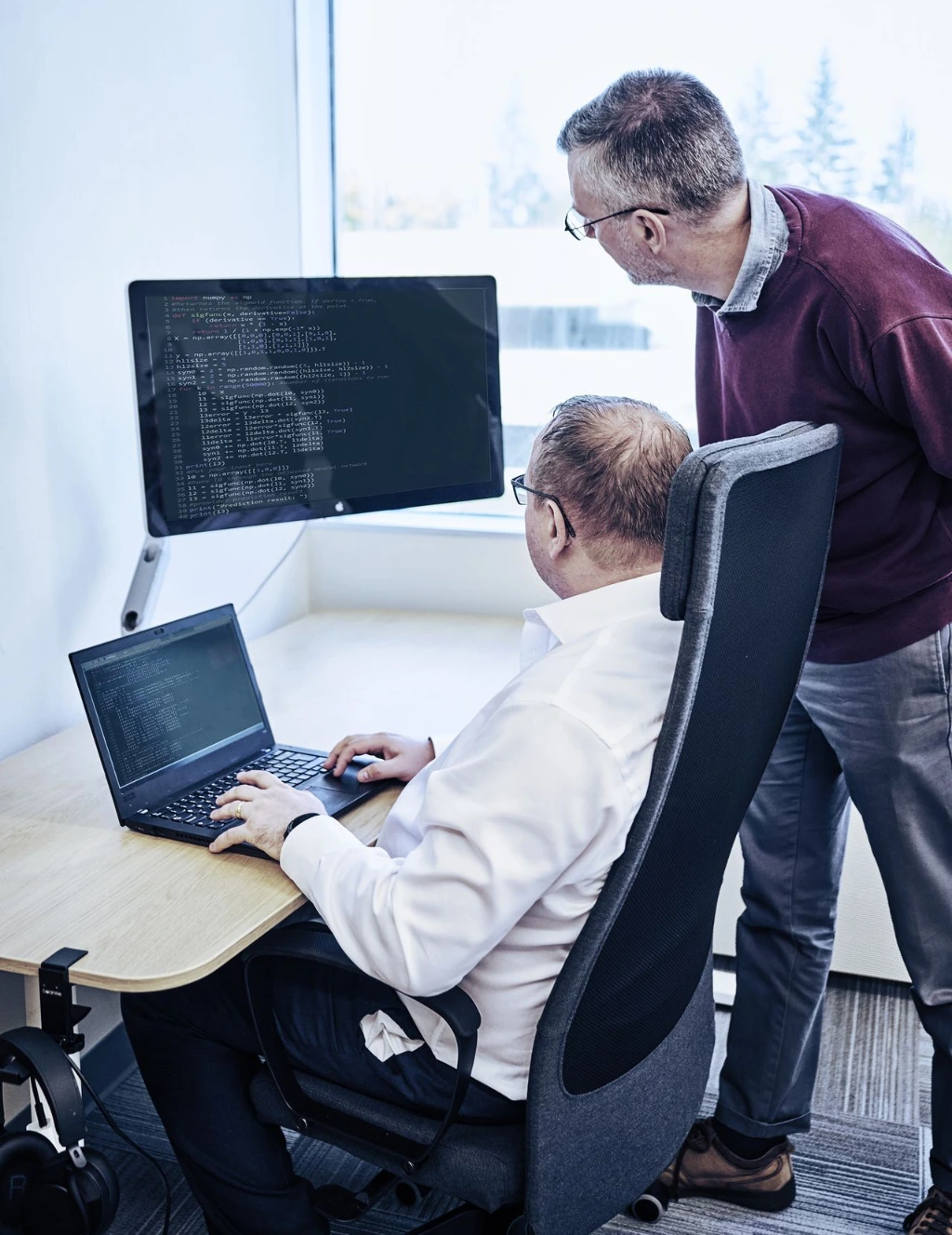 Two men in an office setting, one seated with a laptop and the other standing, looking at a large computer monitor displaying code, collaborating in a bright room with a window showing trees outside.