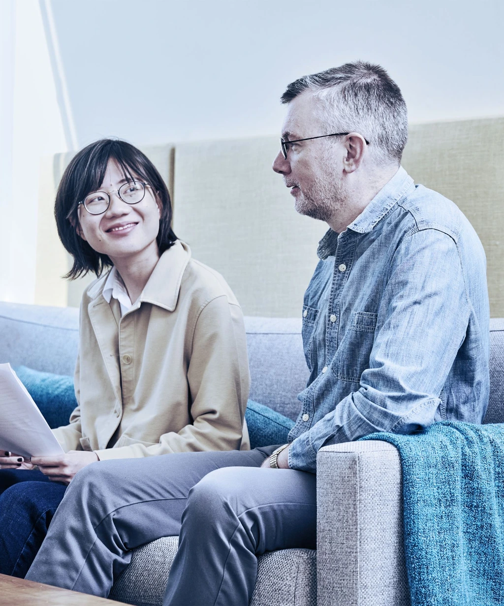 Two people sitting on a couch, one with dark hair and glasses holding papers, the other with gray hair and glasses wearing a denim shirt, engaged in a conversation in a well-lit room with a window and beige wall panels.