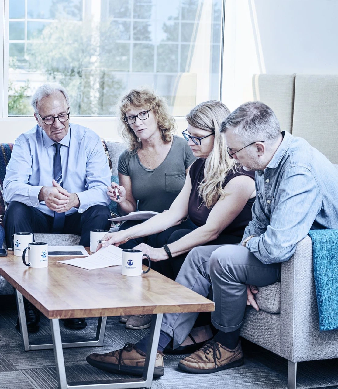 Diverse group of adults sitting on couches and engaged in discussion around a wooden coffee table with mugs and papers in a modern room with large windows and trees visible outside.