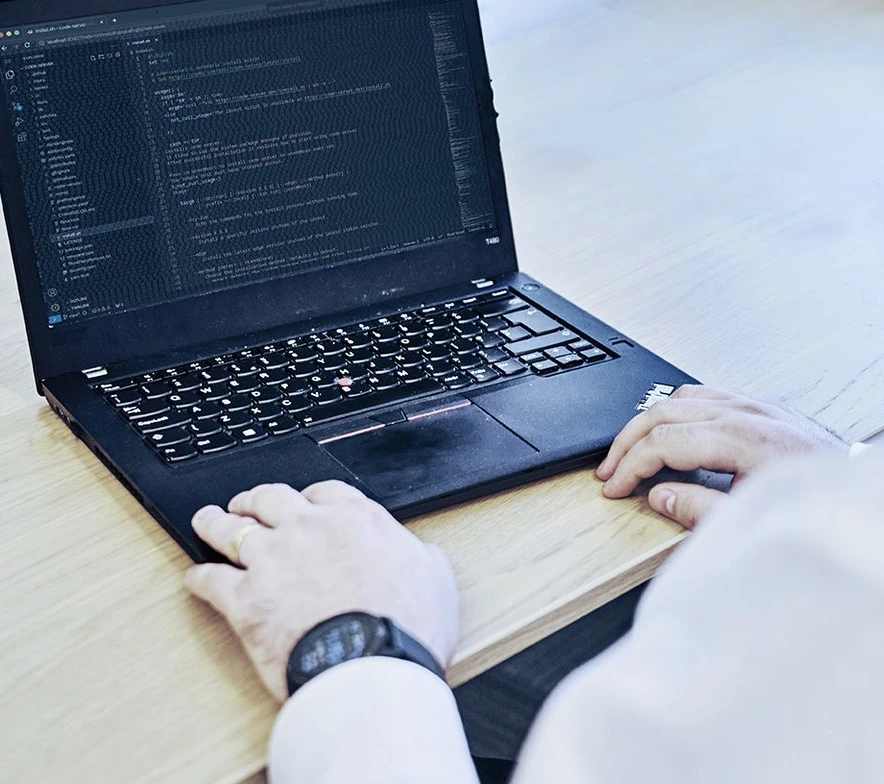 Man in white shirt working on black laptop with code on screen, sitting at light wood desk with Apple monitor above, wearing glasses and watch, in a clean and minimalist office space with white walls and window.