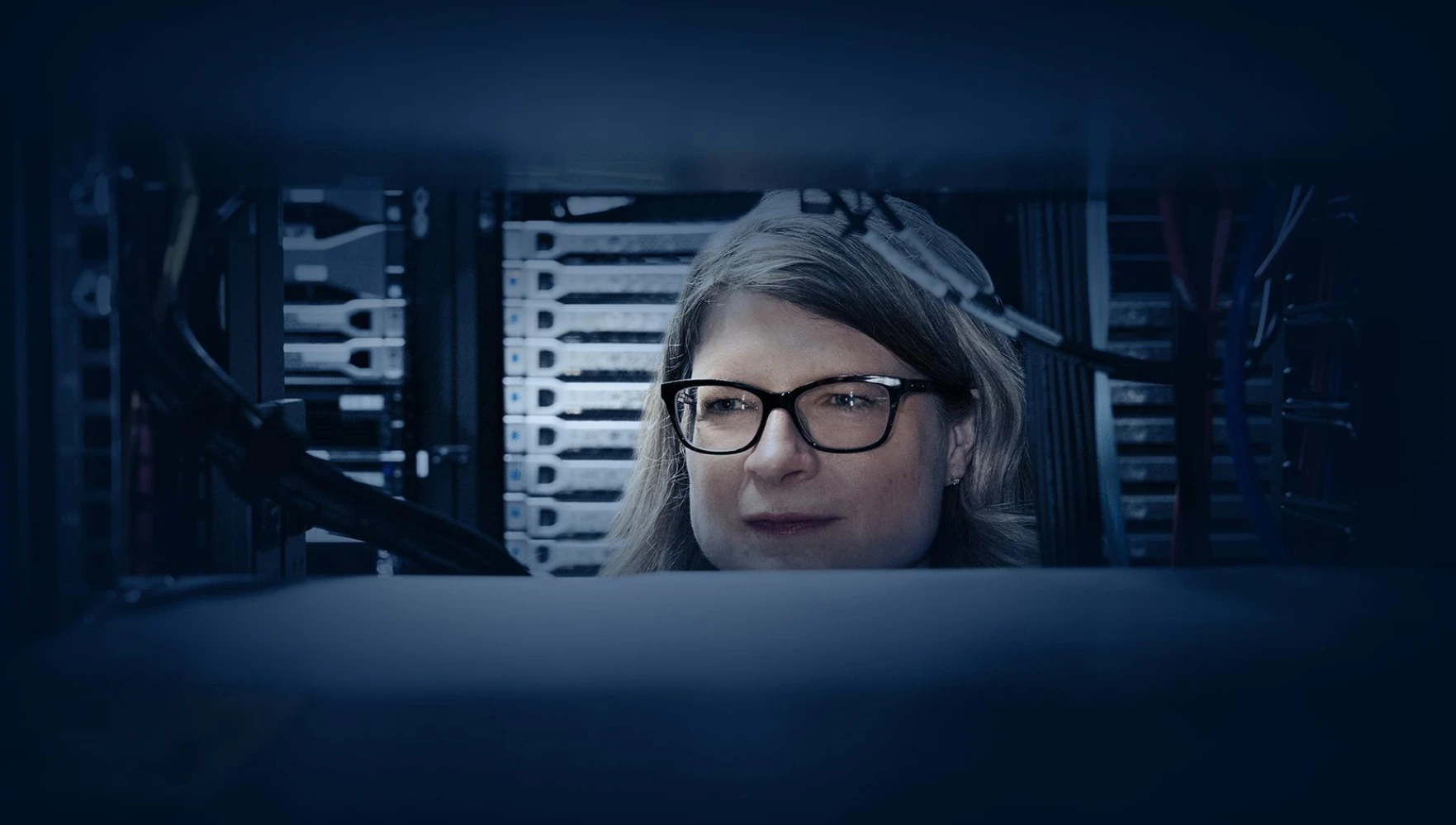 Woman with glasses working on computer in server room, surrounded by rows of computer servers and cables, focused on screen.