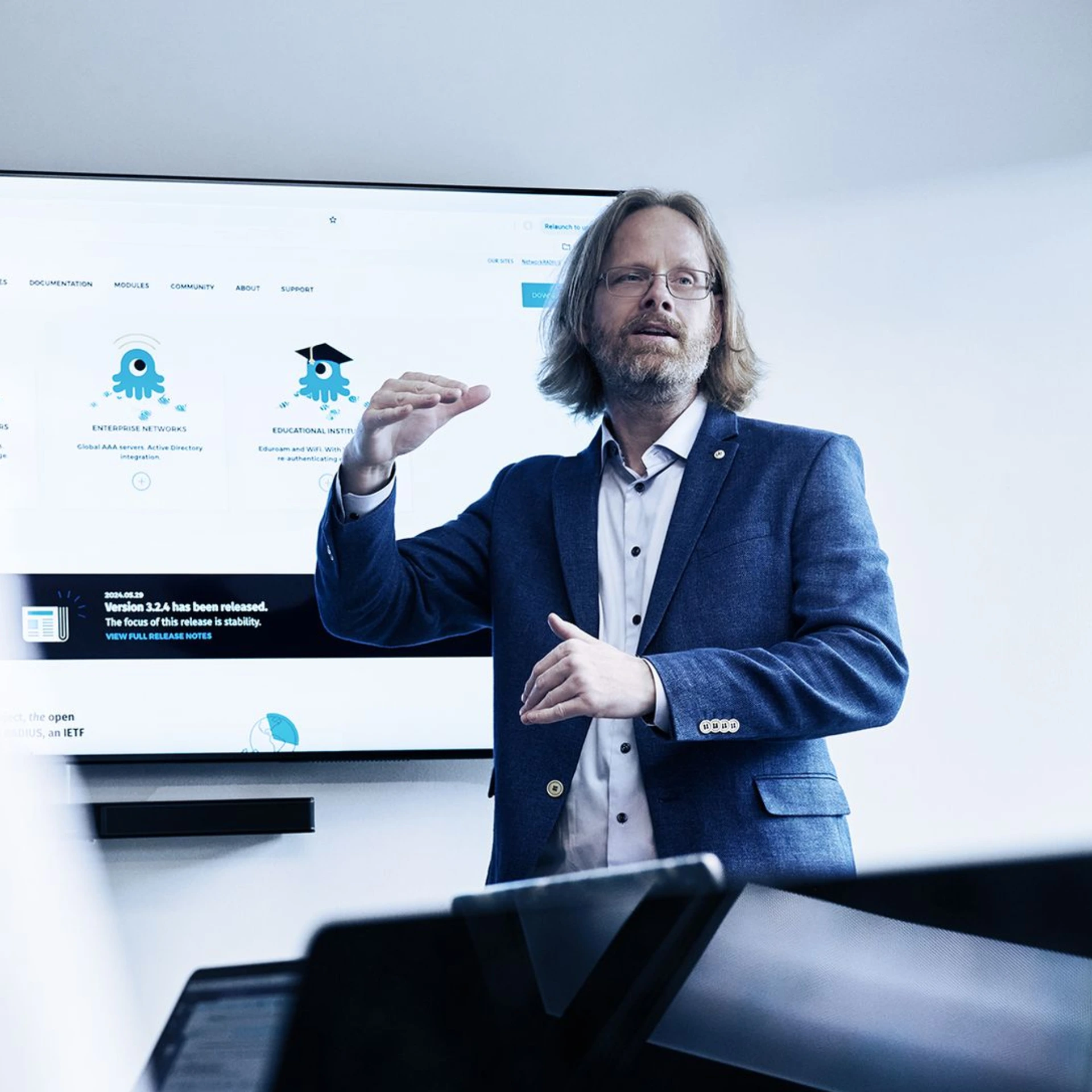 Businessman in blue suit presenting to audience in front of large screen display with educational icons and text, gesturing with hands in professional setting.
