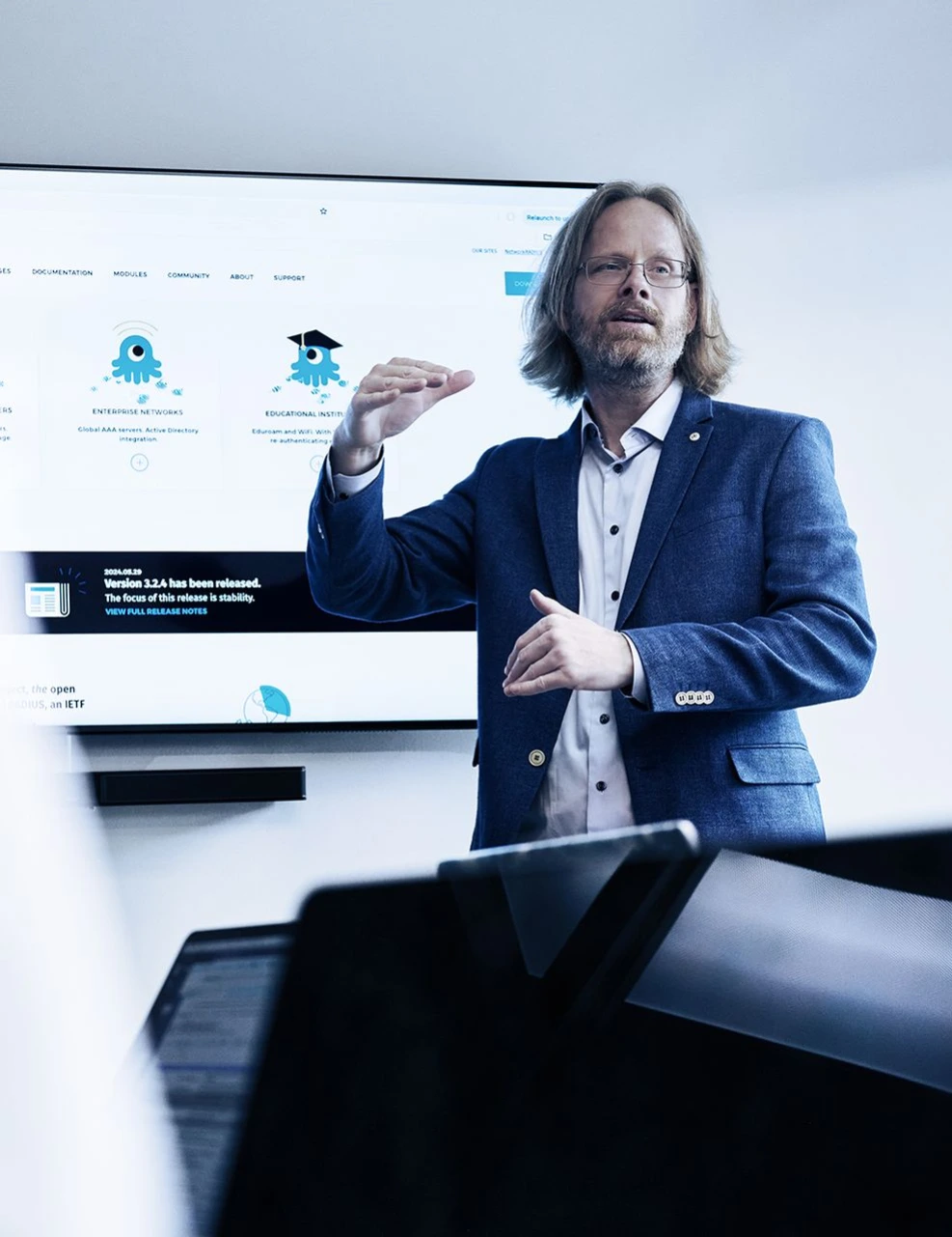 Businessman in blue suit presenting to audience in front of large screen display with educational icons and text, gesturing with hands in professional setting.