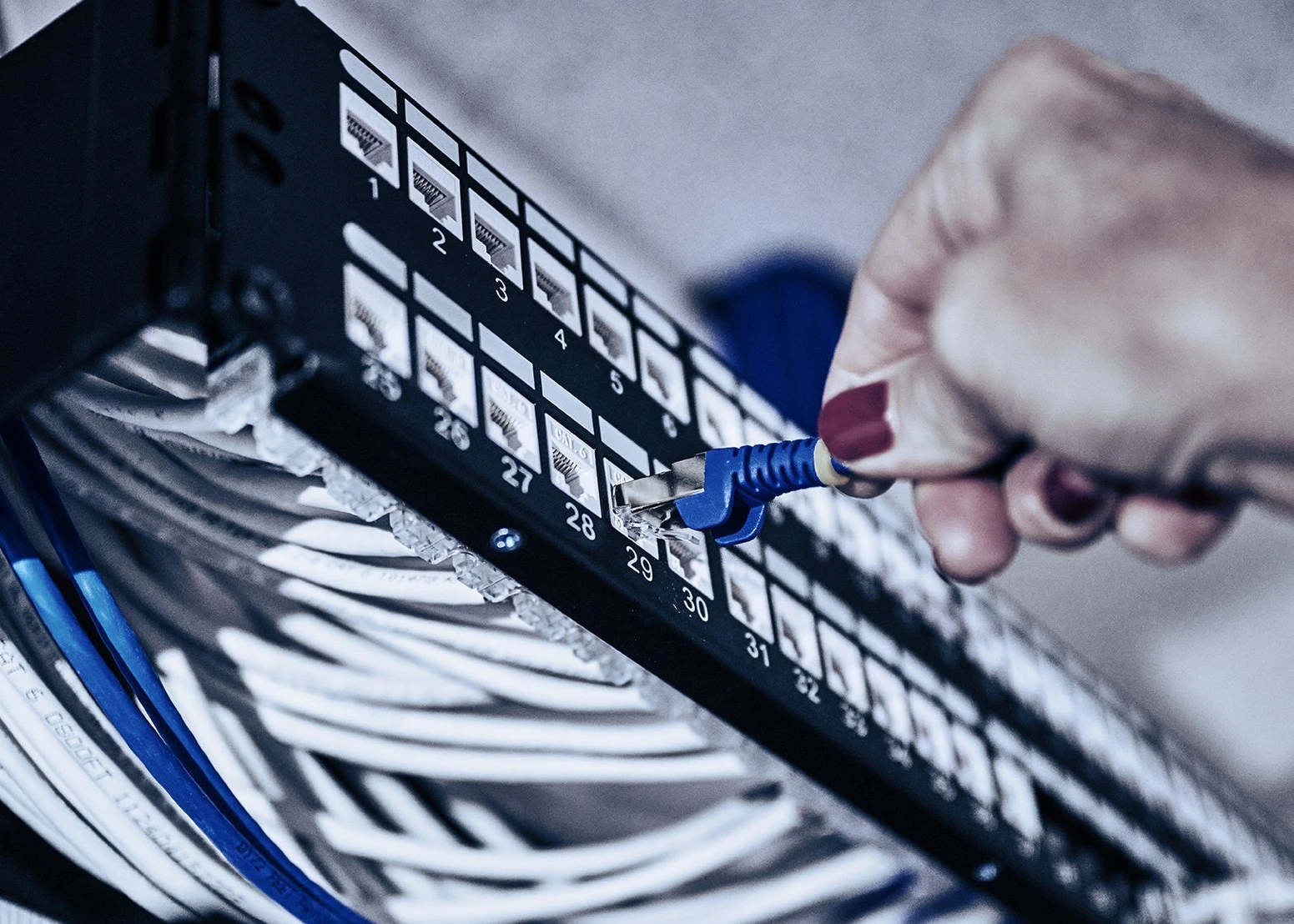 Network switch with cables, hand inserting blue ethernet cable into numbered port. Multiple white and blue wires connected to black patch panel, secure internet connection setup.